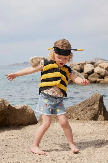 Dziecko w stroju pszczółki z czarnymi i żółtymi paskami z opaską z czułkami i skrzydłami na plaży. Kid’s Concept Strój pszczółki