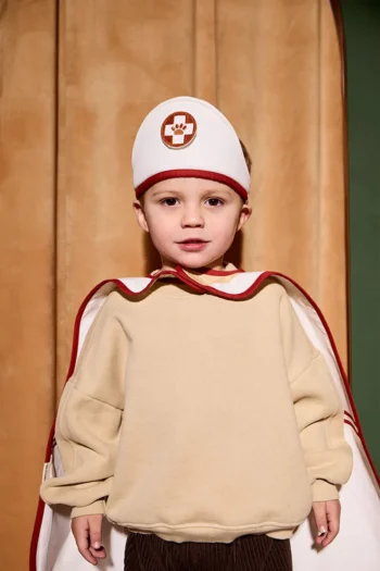 Dziecko w beżowej bluzie i białej czapce z czerwonym paskiem oraz pelerynie kostium doktora dla dziecka. Kid’s Concept Strój doktora