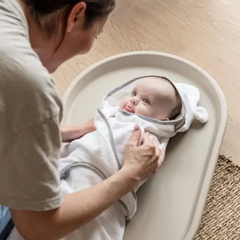 Mata do przewijania z pianki w kolorze taupe niemowlę owinięte w biały ręcznik kąpielowy z uszkami na przewijaku na komodę. Shnuggle Mata do Przewijania z Pianki Squishy Taupe