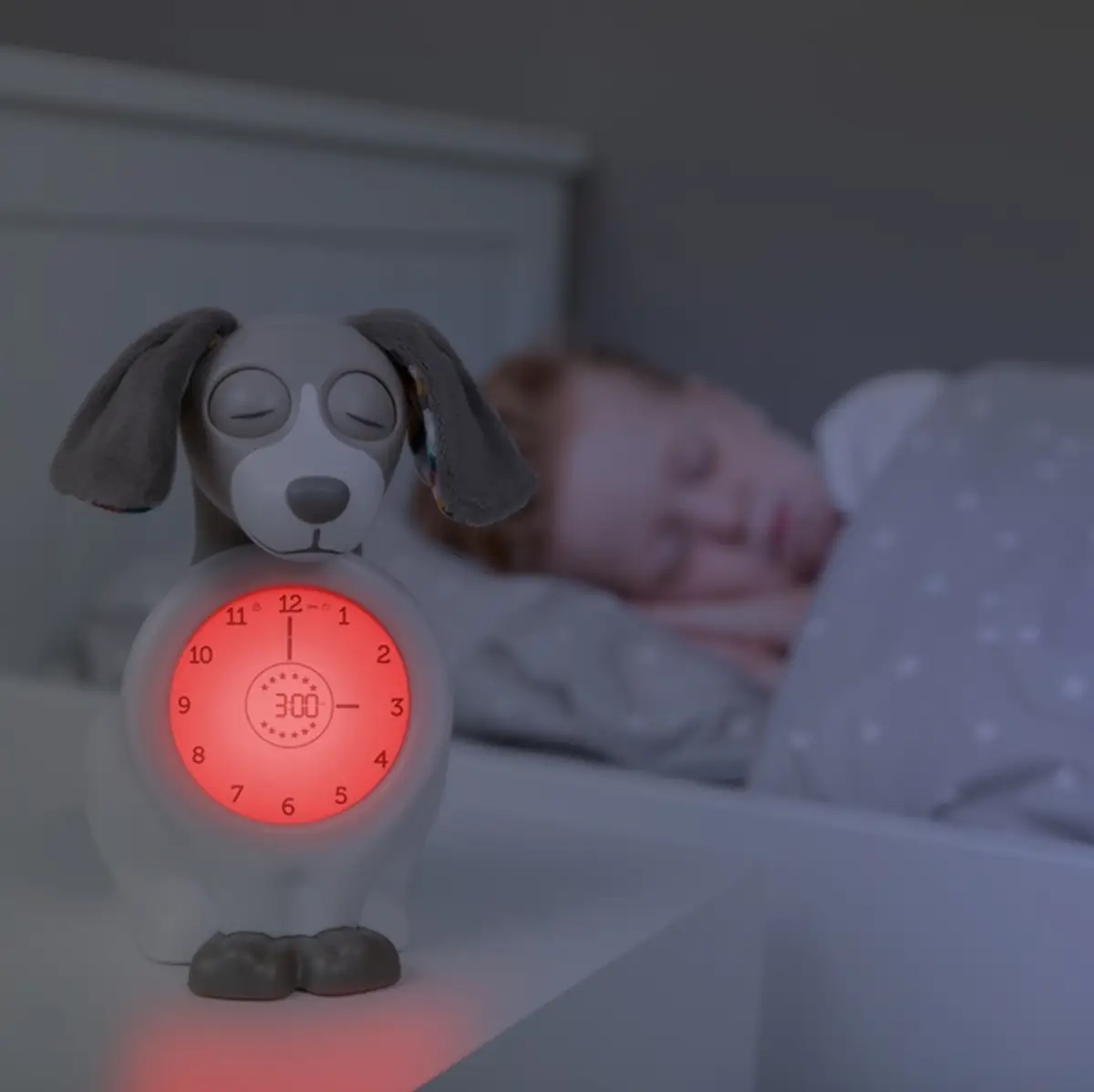 Budzik i trener snu w kształcie psa w kolorze taupe dla dziecka na nocnym stoliku obok śpiącego dziecka. Zazu DAVY Budzik i Trener Snu Pies Taupe