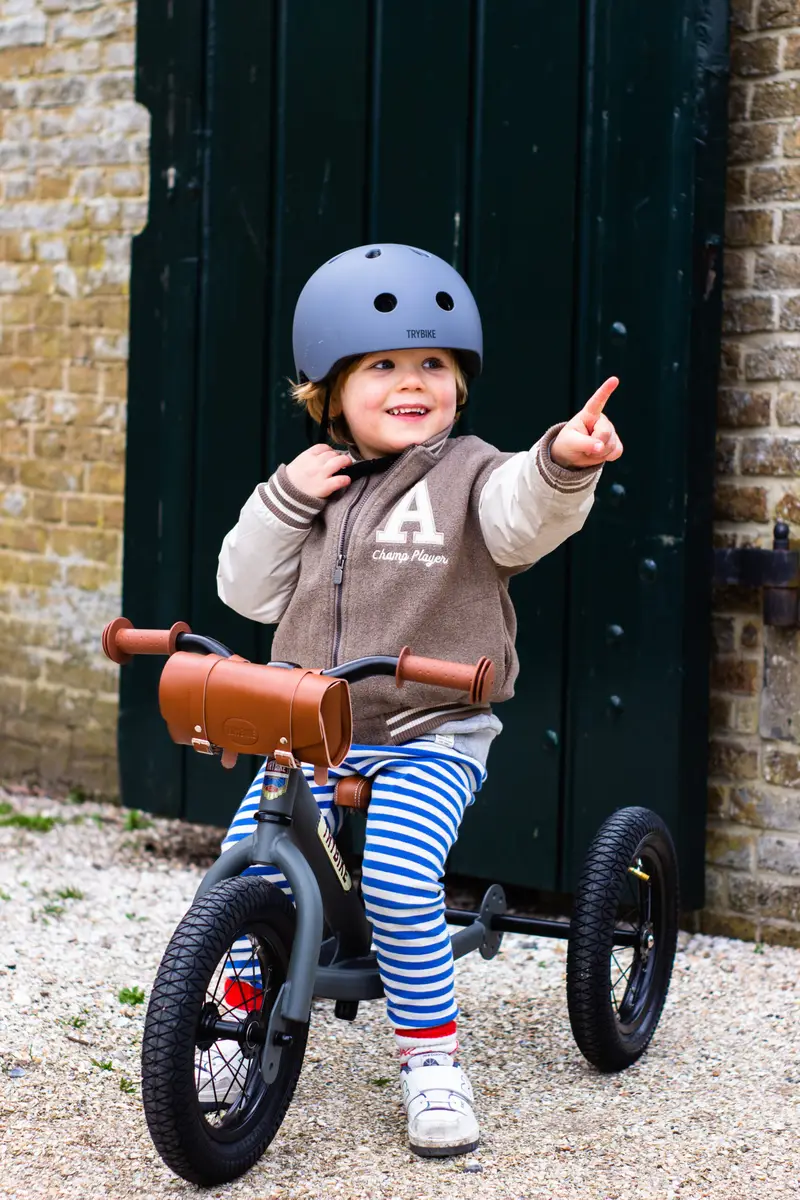Dziecko na rowerku biegowym w szarym kasku dziecięcym XS ubrany w bluzę i spodnie w paski. CoandCo Kask dziecięcy na rower rolki deskorolkę hulajnogę szary XS Trybike