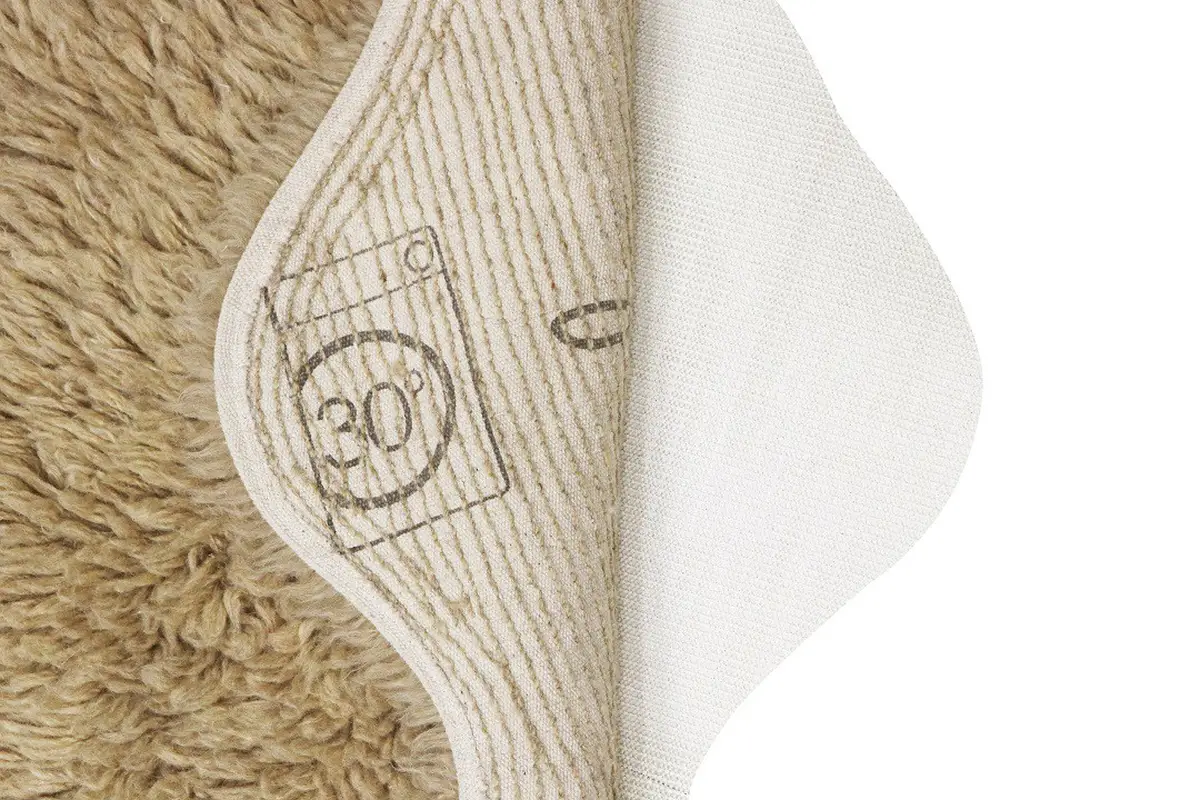 Fragment beżowego dywanu wełnianego z widoczną metką pielęgnacyjną i białą podkładką antypoślizgową. Lorena Canals Dywan wełniany Woolly Beige 75x110cm