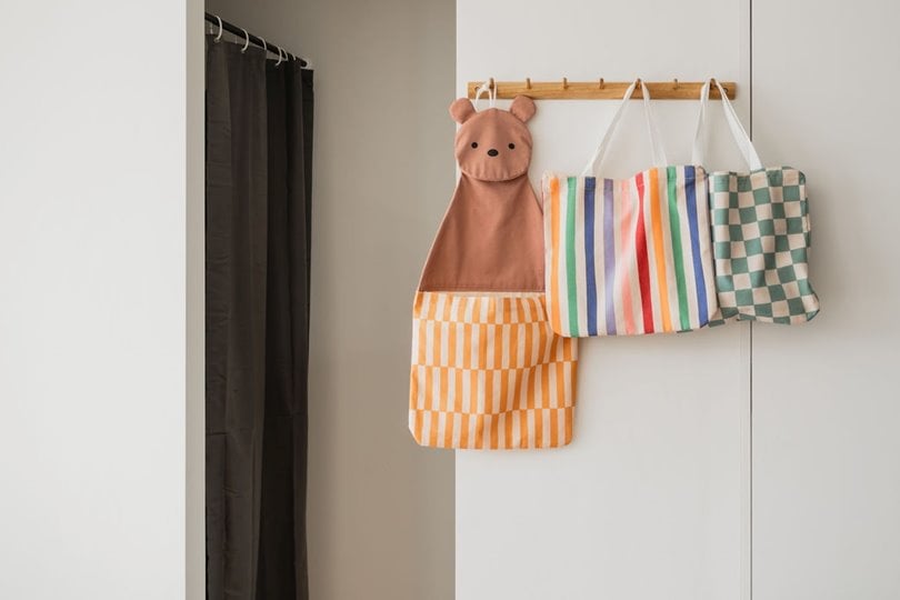 Organizer ścienny i torba dla dzieci w kolorowe paski i kratkę oraz w kształcie zwierzątka na drewnianych haczykach na białej ścianie. Play&Go TOTO Organizer Ścienny i Torba dla Dzieci 2w1 Jednorożec