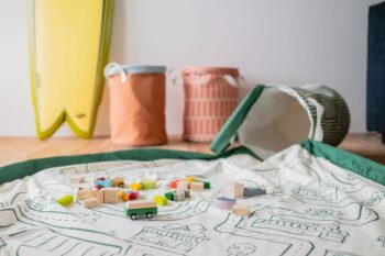 Mata do zabawy z nadrukiem mapy miasta w kolorze białym z zielonym obramowaniem z drewnianymi zabawkami i torbą na zabawki w tle. Play&Go SOO Kosz na Zabawki i Mata do Zabawy 2w1 Roadmap