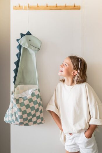 Organizer ścienny i torba dla dzieci w kształcie dinozaura w zielono-białą kratę zawieszony na ścianie obok dziewczynki w białej bluzce i szortach. Play&Go TOTO Organizer Ścienny i Torba dla Dzieci 2w1 Dinozaur