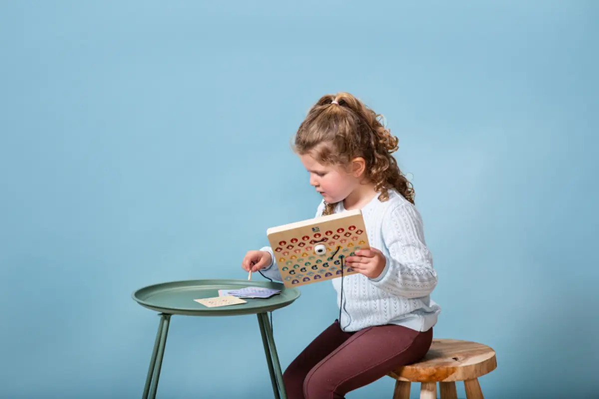 Dziewczynka bawiąca się drewnianą tablicą do kodowania z kartami pracy na zielonym stoliku i drewnianym stołku. BS Toys drewniana tablica do kodowania Przeplatanka z kartami pracy