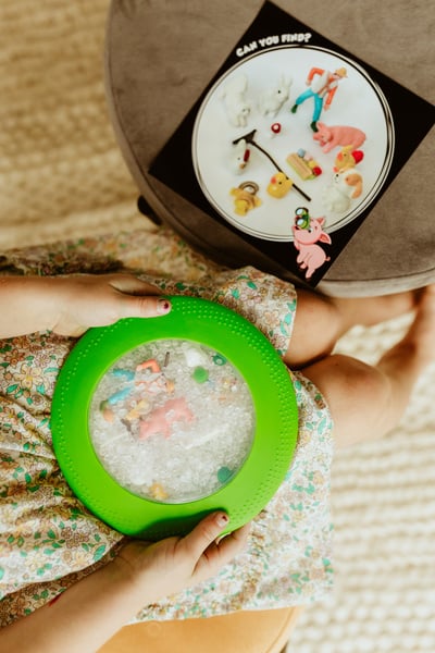 Dziecko trzyma sensoryczny dysk z przezroczystą powierzchnią i kolorową ramką z motywem farmy z figurkami w środku. Jellystone Designs Jellystone Sensoryczny dysk z figurkami A-Ku-Ku! Peekaboo motyw Farma