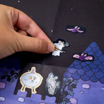 Ręka naklejająca naklejkę z postacią ducha na ciemnym tle z ilustracją domu i nietoperzy dla dzieci. Londji Arts&Crafts Kreatywne Naklejki Enchanted
