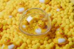 Przezroczysta kula sensoryczna z żółtą kaczuszką i białymi elementami na tle żółtych piłeczek dla niemowlaka. Petit Boum Happy Bubbles kula sensoryczna Kaczuszka