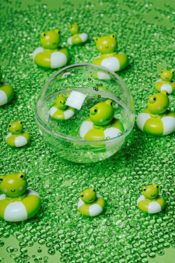 Przezroczysta kula sensoryczna z zieloną żabą na białym kołnierzu do kąpieli dla małego odkrywcy na zielonym tle z zielonymi koralikami. Petit Boum Happy Bubbles kula sensoryczna Żaba