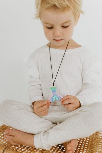 Dziewczynka trzymająca pastelowy naszyjnik z zawieszką w kształcie syreny z silikonu na czarnym sznurku dla dziecka. Jellystone Designs Jellystone Gryzak terapeutyczny antystresowy naszyjnik Syrena