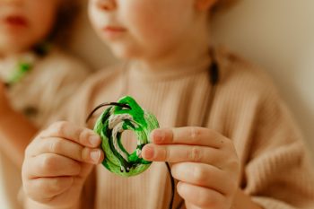 Dziecko trzyma zielony terapeutyczny gryzak naszyjnik w kształcie węża dla niemowlaka. Jellystone Designs Jellystone Gryzak terapeutyczny antystresowy naszyjnik Leśny Wąż