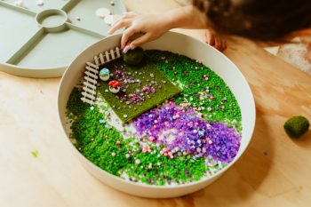 Taca sensoryczna z zielonym i fioletowym materiałem ziarnistym oraz małymi dekoracjami do zabawy dla dziecka. Jellystone Designs Jellystone Zestaw sensoryczny Zaczarowany Ogród wkład do tacki sensorycznej Play Tray