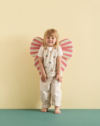 Dziecko w białej koszulce i beżowych spodniach z różowo białymi skrzydełkami w paski dla dziecka. Kid’s Concept Strój kokarda różowa