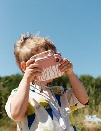 Dziecięcy aparat fotograficzny w kolorze różowym z wbudowaną drukarką trzymany przez dziecko na tle nieba i zieleni. Hoppstar Aparat fotograficzny dla dzieci z drukarką Artist Blush