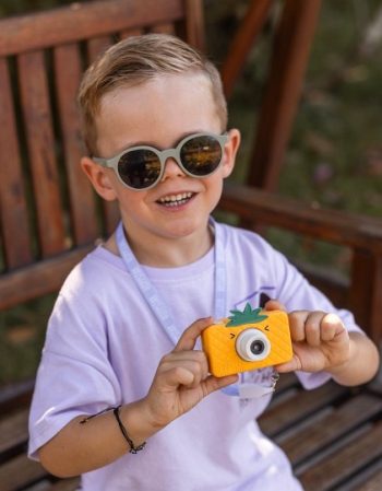Dziecko w okrągłych okularach przeciwsłonecznych trzyma pomarańczowy aparat fotograficzny dla dzieci z motywem ananasa. Hoppstar Aparat fotograficzny dla dzieci Rookie Pina Coolada