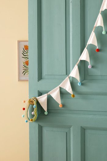 Girlanda urodzinowa z białych trójkątnych flag z kolorowymi pomponami dla dziecka zawieszona na zielonych drzwiach. Kid’s Concept urodzinowa girlanda