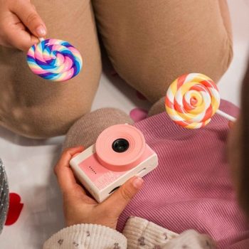 Dziecięcy aparat fotograficzny w kolorze różowo-białym trzymany przez dziecko obok kolorowych lizaków spirali. Hoppstar Aparat fotograficzny dla dzieci Popcorn Sorbet