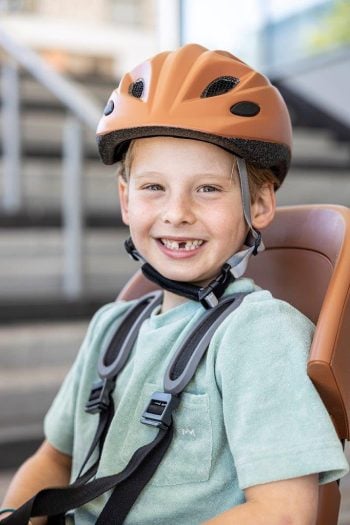 Dziecko w pomarańczowym kasku rowerowym dla dzieci XS 48-52 cm siedzi na foteliku rowerowym. Bobike KASK dla dzieci XS 48-52cm Chocolate Brown