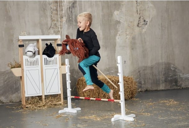 Przeszkoda do skakania dla konia na kiju hobby horse biała z czerwoną poprzeczką na tle stodoły z sianem i dzieckiem skaczącym na koniku na kiju. byAstrup Przeszkoda do skakania dla konia na kiju Hobby Horse