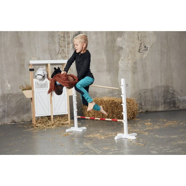 Dziecko skaczące przez przeszkodę na koniu na kiju z brązową grzywą na tle stodoły. byAstrup Koń na kiju Hobby Horse szary z szarą grzywą