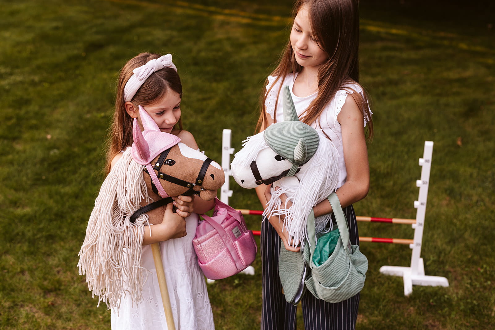 Dwie dziewczynki bawiące się konikami na patyku z różową podkładką pod siodło i bonetką dla hobby horse konia na zewnątrz na trawie. byAstrup Podkładka pod siodło i bonetka dla Hobby Horse konia, różowa