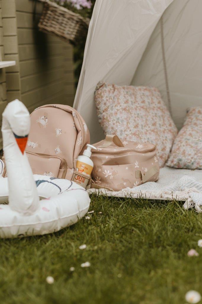 Beżowa torba termiczna na lunch z nadrukiem kwiatów na trawie obok poduszki i dmuchanego koła w kształcie łabędzia. Vanilla Copenhagen Torba termiczna Cooler Bag NEO LILY Oak