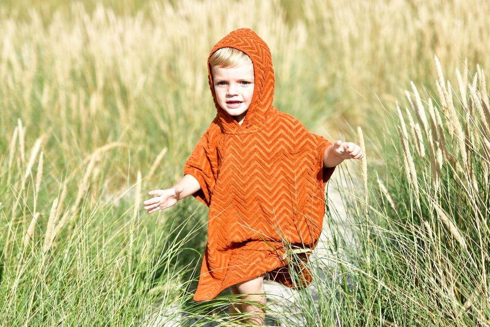 Pomarańczowe ponczo kąpielowe z kapturem w zygzak dla dziecka na tle trawy. Filibabba Poncho Zigzag Rust