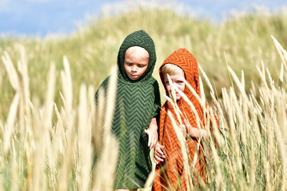 Dwa dzieci w kąpielowych ponczach z kapturem w zygzakowy wzór zielonym i rudym na polu zboża. Filibabba Poncho Zigzag Rust