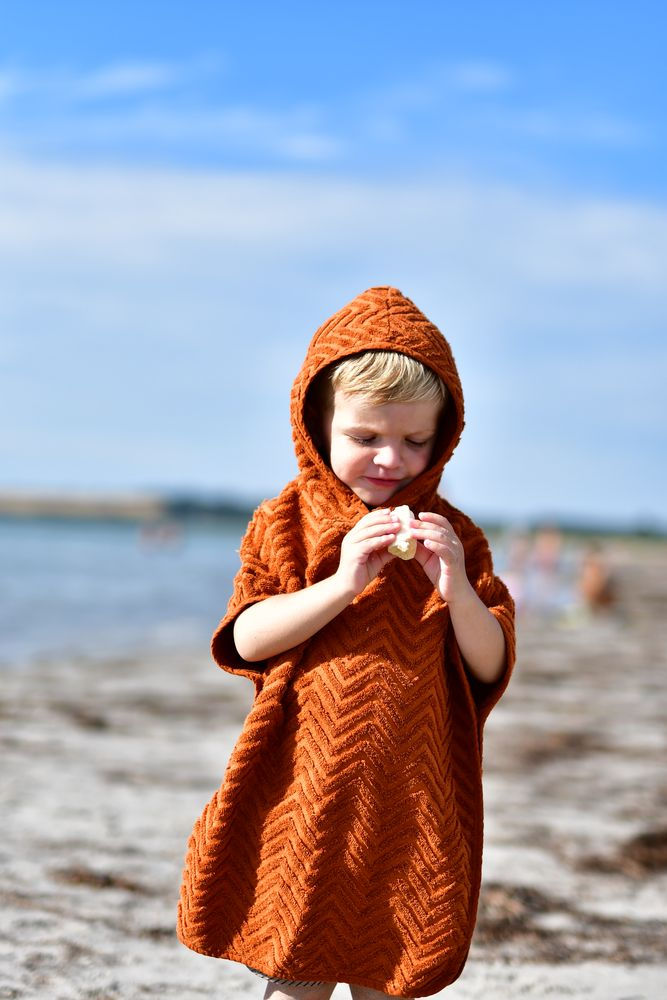 Dziecko na plaży w pomarańczowym ponczo kąpielowym z kapturem z wzorem zygzakowym. Filibabba Poncho Zigzag Rust