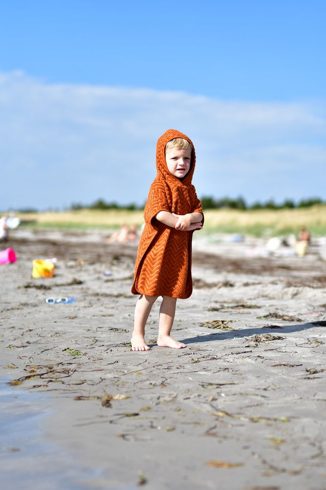 Dziecko na plaży w pomarańczowym ponczo kąpielowym z kapturem z wzorem zygzakowym. Filibabba Poncho Zigzag Rust
