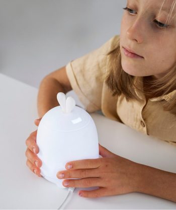 Biała lampka nocna w kształcie królika z uszami trzymana przez dziecko na białym stole. Rabbit&Friends Królik Dyfuzor do Olejków Lampka Biała