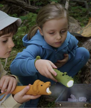 Dzieci bawiące się latarkami w kształcie krokodyli w kolorze zielonym i pomarańczowym na zewnątrz w lesie. Rabbit&Friends Latarka Krokodyl CROCO