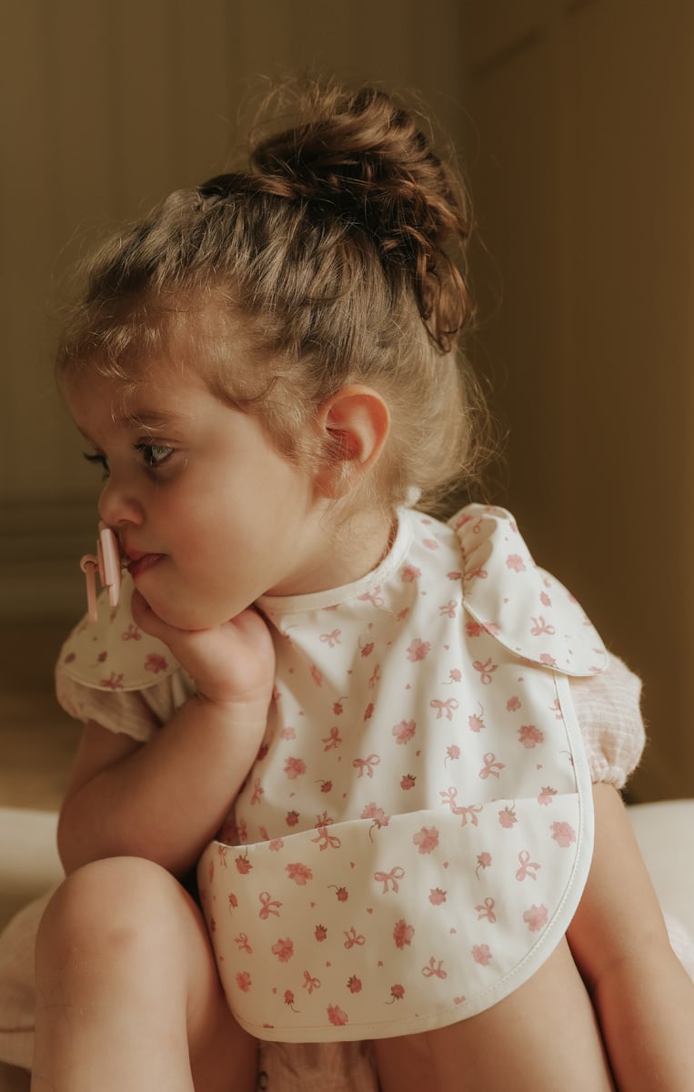Materiałowy śliniak dla dziecka w jasnym kolorze z różowym wzorem kwiatowym i kokardek. Elodie Details śliniak petit river rose