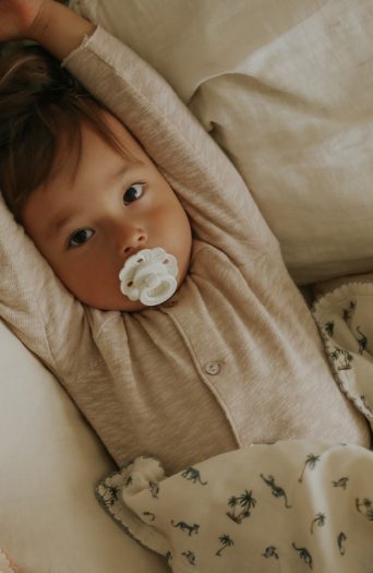 Niemowlę z lateksowym smoczkiem w kolorze białym leżące na łóżku z jasną pościelą dla niemowląt. Elodie Details smoczek lateksowy Binky Bloom 3 m+ valinna white