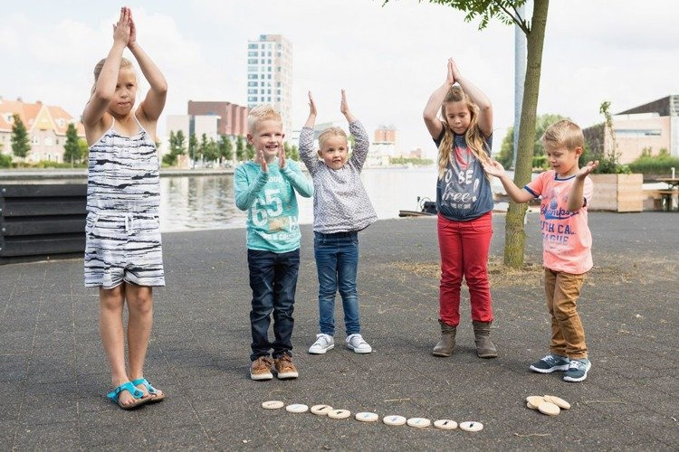 Pięcioro dzieci na zewnątrz na placu zabaw bawiących się w grę naśladuj ruch z drewnianymi elementami na ziemi. BS Toys Gra Naśladuj Ruch