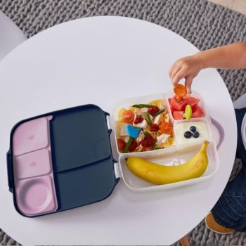 Biało granatowa śniadaniówka z przegródkami z jedzeniem i dziecko wkładające pokrojone owoce dla dziecka. b.box Lunchbox Indigo Rose