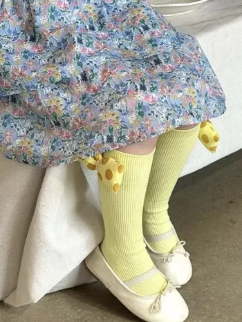 Żółte prążkowane podkolanówki z 3D kokardą dla dziecka na nogach w białych balerinach i sukience w kwiaty. Fig&Pear Podkolanówki prążkowane z żółtą kokardą 3D