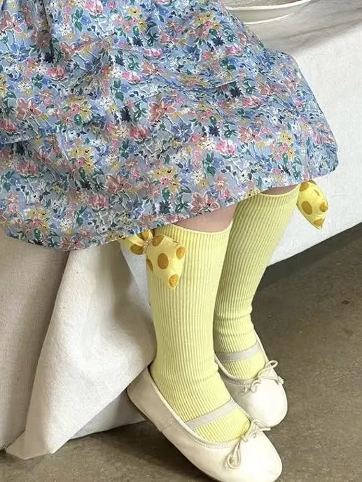 Żółte prążkowane podkolanówki z 3D kokardą dla dziecka na nogach w białych balerinach i sukience w kwiaty. Fig&Pear Podkolanówki prążkowane z żółtą kokardą 3D