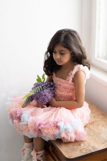 Tiulowa sukienka dziecięca w kolorze koralowym z falbanami i pastelowymi akcentami dziewczynka siedzi z bukietem kwiatów. Fig&Pear Sukienka tiulowa Baby Coral Rainbow