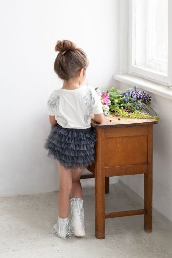 Dziewczynka w szarych falbaniastych bloomersach i białej bluzce stoi przy drewnianym stoliku z kwiatami. Fig&Pear Bloomersy Midnight Grey