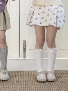 Białe podkolanówki z kokardkami dla dziewczynki na nogach w białej spódnicy z kwiatowym wzorem. Fig&Pear Skarpetki w złamanej bieli z kokardkami