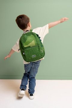 Zielony plecak w kształcie aligatora dla dziecka na plecach chłopca w jasnej koszulce i dżinsach na tle zielonej ściany. Trixie Plecak Aligator Baby