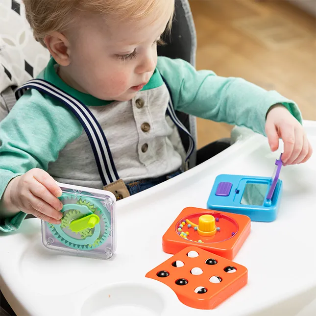 Dziecko bawi się sensorycznym kwadratem z lusterkiem i kolorowymi elementami na białym stole. Fat Brain Toy Co. PlayTab Sensoryczny Kwadrat Lusterko