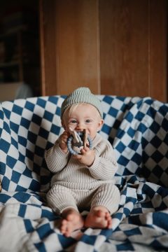 Niemowlę w beżowym ubranku i czapce trzymające silikonowy gryzak bransoletkę z zawieszkami w kolorze szarym siedząc na niebiesko-białym kocu. Mushie Gryzak Silikonowy Bransoletka z Zawieszkami Space