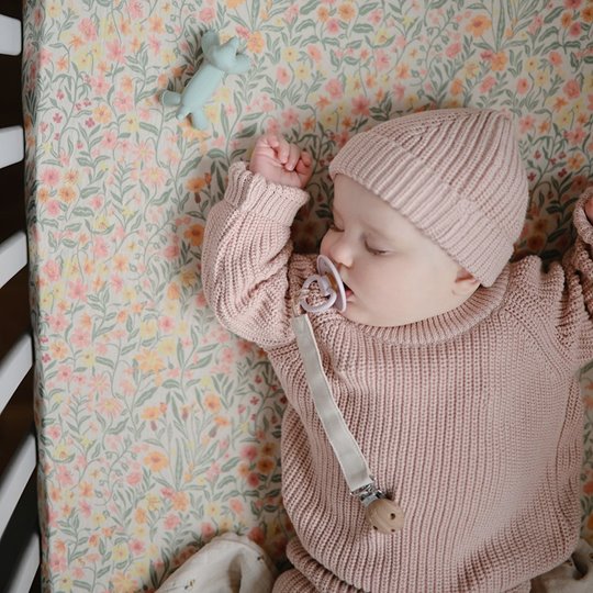Niemowlę śpiące w różowym sweterku i czapce z silikonowym gryzakiem pieskiem na smoczku na łóżeczku z kolorową pościelą. Mushie Gryzak silikonowy figurka Piesek