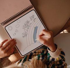 Dziecko koloruje wodną kolorowankę z wróżkami i tęczą na spiralnym bloku. Mushie Wodna Kolorowanka Wielokrotnego Użytku Fairies / Wróżki