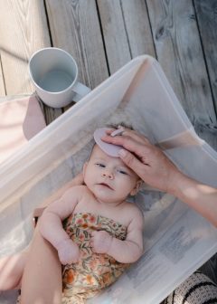 Niemowlę w wanience z ręką trzymającą różową szczotkę do ciemieniuchy dla niemowląt. Mushie Szczotka na Ciemieniuchę dla Niemowląt Rose / Soft Lilac
