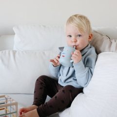 Dziecko pijące z niebieskiego kubka ze słomką z motywem retro samochodów na białej kanapie. Mushie Kubek ze słomką 175ml Retro Cars 175 ml