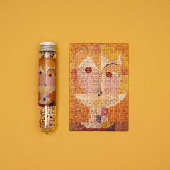 Puzzle kieszonkowe z obrazem twarzy w stylu Paula Klee w tubie i ułożone na żółtym tle. Londji Puzzle mikro menzurka Paul Klee Senecio
